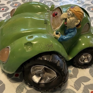 May include: A green cartoon-style car figurine with oversized black tires and silver rims. A character with blonde hair and a blue shirt is driving the vehicle. The car features a small red ladybug detail on the windshield and textured surface markings.