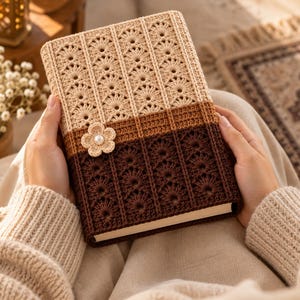 May include: A rectangular book cover featuring a two-tone crochet design with a light beige upper section and a dark brown lower section. The cover displays a repeating fan-like stitch pattern and is adorned with a small crocheted flower with a pearl center.