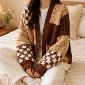 May include: A long-sleeve crochet cardigan featuring a patchwork design in shades of dark brown, tan, and cream. The garment includes panels with a white floral motif on the sleeves and upper chest, finished with ribbed cuffs and a front opening.