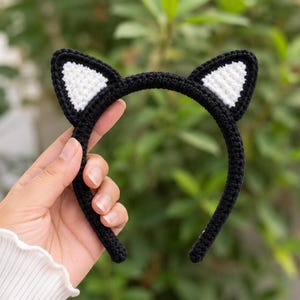 May include: A black crochet headband featuring two pointed cat ears with white triangular centres. The headband is constructed with a dense, textured yarn stitch pattern, creating a solid black frame with contrasting white inner ear details.