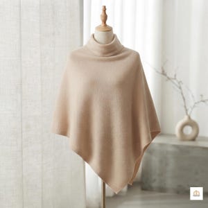 May include: A beige knit poncho featuring a high turtleneck collar and a soft, textured surface. The garment is displayed on a mannequin, showcasing an asymmetrical hemline and a simple, solid-coloured design with ribbed detailing at the edges.