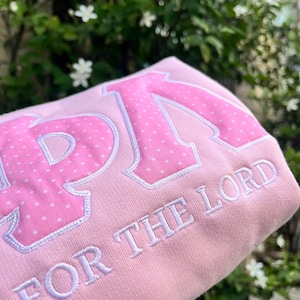 May include: A light pink sweatshirt featuring large, appliqued Greek letters in a pink fabric with a white polka dot pattern. Below the letters, the phrase FOR THE LORD is embroidered in white thread with a clean, sans-serif font style.