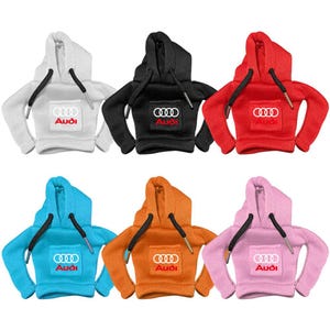 May include: A collection of six miniature hoodies in white, black, red, blue, orange, and pink. Each hoodie features a hood, long sleeves, black drawstrings, and a front pocket with the white Audi logo and text printed on the centre of the pocket.