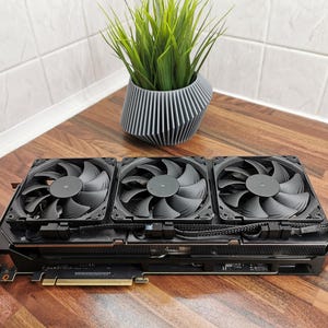 May include: A black ASUS graphics card featuring three cooling fans mounted on top of a heatsink, with visible black cabling connecting the fans, a gold-plated PCIe connector, and a metallic backplate visible along the bottom edge of the component.