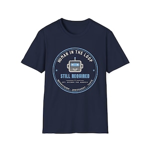 May include: A navy blue short-sleeve t-shirt featuring a circular graphic with a robot icon and the text "HUMAN IN THE LOOP STILL REQUIRED" along with "SUPERVISION", "JUDGEMENT", and "TASTE" printed in white and light blue on the chest.