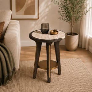 Puede incluir: Una mesa auxiliar redonda con una superficie de mármol claro y una estructura de madera oscura con detalles verticales dorados en las patas. La mesa incluye un estante circular inferior y sostiene un vaso de vidrio oscuro y un cuenco metálico dorado.