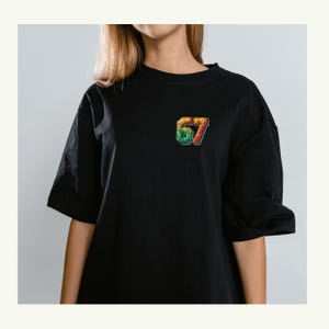 May include: Black t-shirt with a colorful, pixelated "67" graphic on the chest. The short-sleeved shirt has a relaxed fit and a crew neck. The graphic features shades of green, yellow, red, and blue.