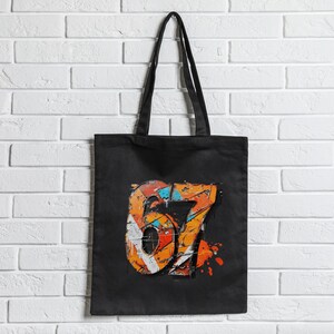 May include: Black tote bag with a colorful graphic of the number 67. The design features orange, white, blue, and red accents with a distressed, graffiti-style aesthetic. The bag has long black handles.