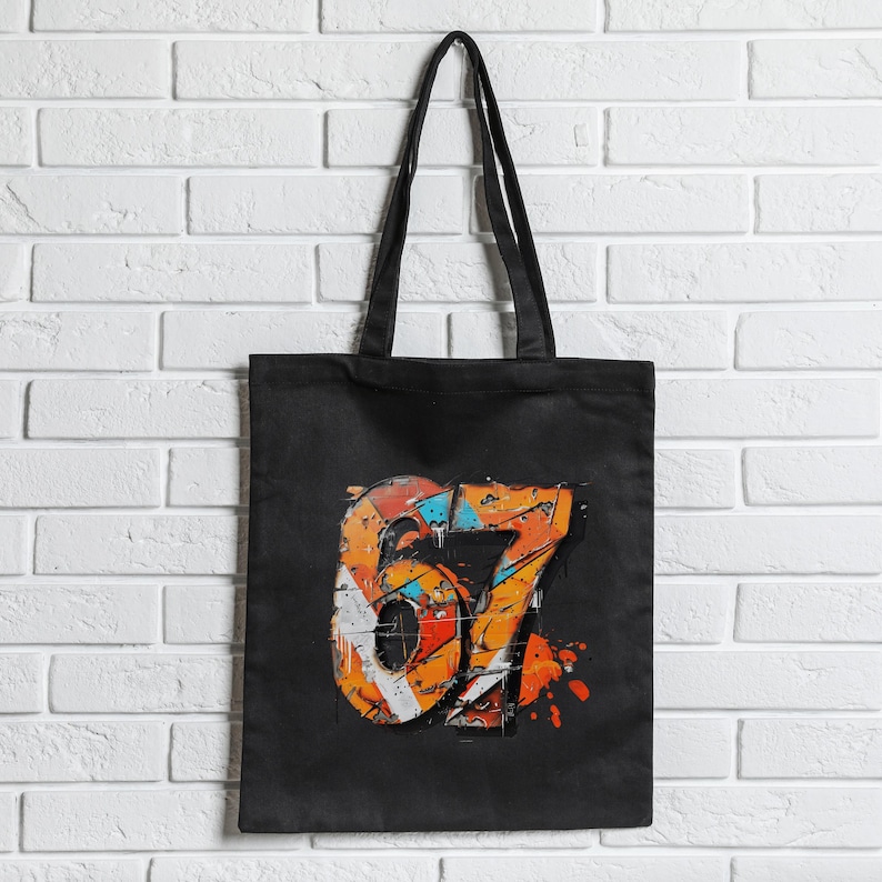 May include: Black tote bag with a colorful graphic of the number 67. The design features orange, white, blue, and red accents with a distressed, graffiti-style aesthetic. The bag has long black handles.