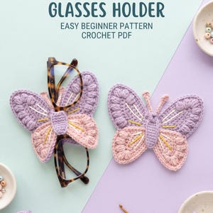 May include: Two crocheted butterfly-shaped glasses holders in lavender and light pink with yellow and white embroidery details. One butterfly holds tortoise-shell glasses. Text reads: BUTTERFLY GLASSES HOLDER EASY BEGINNER PATTERN CROCHET PDF.