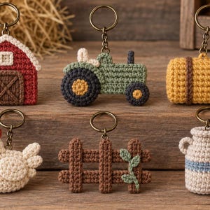 Op de afbeelding: Een set van zes gehaakte sleutelhangers met boerderijthema, waaronder een rode schuur, een groene tractor, een gele hooibaal, een witte kip, een bruin houten hek met groene bladeren en een beige melkkan, bevestigd aan bronzen metalen ringen.