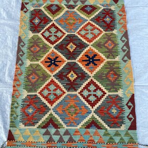 May include: A rectangular woven rug featuring a repeating diamond pattern in shades of teal, red, orange, green, and dark gray. The design includes intricate geometric motifs within each diamond, finished with a short fringe at both ends.