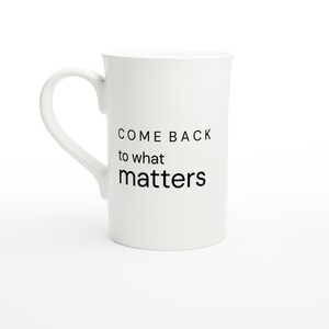 May include: A white ceramic mug with a curved handle featuring the black printed text COME BACK to what matters. The mug has a smooth, cylindrical body and a clean, minimalist design suitable for holding hot or cold beverages.