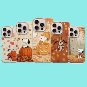 May include: Five clear smartphone cases featuring Snoopy and Woodstock in various autumn scenes. Designs include Snoopy on a pumpkin, inside a jack-o'-lantern, playing in fallen leaves, sitting by a doghouse, and wearing a scarf in a forest.