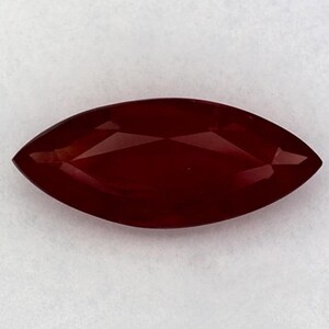 Puede incluir: Una piedra preciosa de color rojo intenso con talla marquesa y superficie facetada. La piedra presenta una forma ovalada alargada y puntiaguda con bordes simétricos y un acabado pulido que refleja la luz sobre su estructura cristalina.