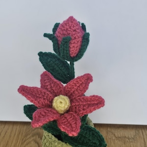 Op de afbeelding: Een gehaakt bloemstuk met een roze knop en een volledig bloeiende roze bloem met een geel hart. De plant heeft donkergroene bladeren en staat in een textuurrijke, licht olijfgroene gehaakte pot, wat zorgt voor een handgemaakte botanische display.
