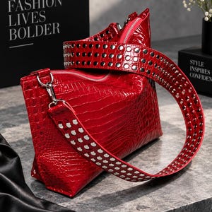 Puede incluir: Un bolso de mano rojo brillante con textura de cocodrilo y acabado lustroso, que cuenta con una correa ancha y extraíble decorada con filas de tachuelas metálicas plateadas. El bolso tiene cierre de cremallera y herrajes plateados.
