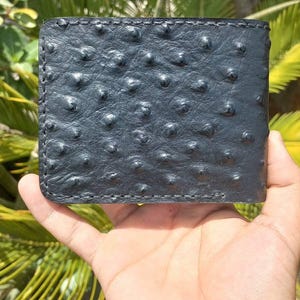 Puede incluir: Una cartera negra plegable hecha de cuero de avestruz texturizado que presenta un patrón de plumas distintivo en toda la superficie. La cartera está construida con costuras visibles a lo largo de los bordes, mostrando un acabado mate oscuro y forma rectangular.