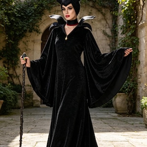 May include: A floor-length black velvet gown with wide bell sleeves and a high, structured collar. The outfit includes a black headpiece with two curved horns, a red choker, and a long, dark, twisted staff with a decorative top ornament.