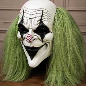 Puede incluir: Una máscara de payaso de cabeza completa con un rostro blanco pálido, marcas negras profundas en los ojos, una amplia sonrisa negra y una pequeña nariz en espiral roja y blanca. La máscara está enmarcada por largo cabello sintético verde claro.