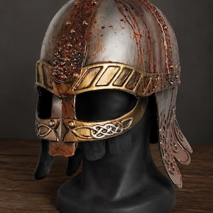 May include: A metallic helmet featuring a silver dome with red splatters, a gold-toned brow band, and a nose guard with intricate silver knotwork patterns. The helmet includes side cheek plates and a central ridge adorned with small rivets.
