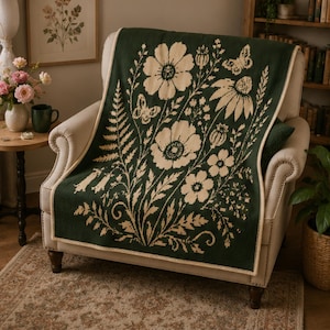 May include: A dark green knitted throw blanket featuring a cream-coloured botanical pattern of large flowers, ferns, and butterflies. The rectangular blanket has a finished cream-coloured edge and is draped over a light-coloured upholstered armchair.