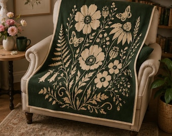 Puede incluir: Una manta de punto de color verde oscuro con un patrón botánico de color crema que muestra flores grandes, helechos y mariposas. La manta rectangular tiene un borde acabado en color crema y está colocada sobre un sillón tapizado claro.
