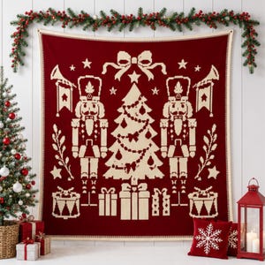 May include: A deep red knit throw blanket featuring a cream-colored pattern of two nutcrackers flanking a central Christmas tree, topped with a bow and surrounded by stars, trumpets, and gift boxes, finished with a cream-colored border.