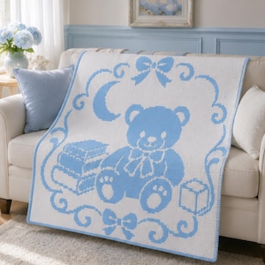 May include: A white knitted blanket featuring a light blue pixelated design of a teddy bear sitting next to a stack of books and a block. The pattern is framed by a decorative border with a crescent moon and two bows at the top and bottom.