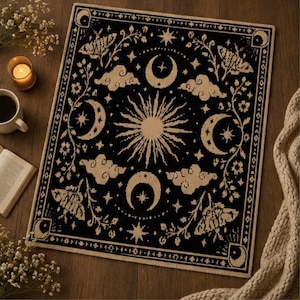 Puede incluir: Una alfombra rectangular tejida en negro y crema con un motivo central de sol radiante rodeado de lunas crecientes, nubes estilizadas, estrellas e ilustraciones de polillas. El dise&ntilde;o est&aacute; enmarcado por un borde decorativo con lunas.