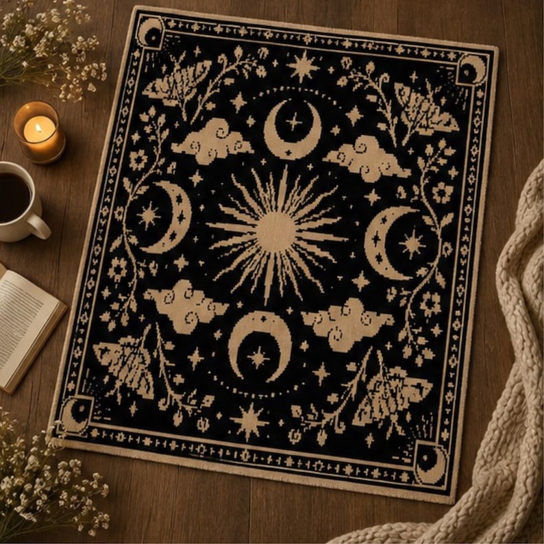 Puede incluir: Una alfombra rectangular tejida en negro y crema con un motivo central de sol radiante rodeado de lunas crecientes, nubes estilizadas, estrellas e ilustraciones de polillas. El dise&ntilde;o est&aacute; enmarcado por un borde decorativo con lunas.