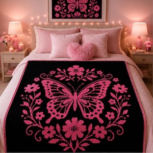 Puede incluir: Una manta negra con un gráfico grande de una mariposa rosa en el centro, rodeada por un patrón circular de flores y hojas rosas. La manta tiene un borde rosa a juego y se muestra sobre una cama con almohadas rosas y un cuadro a juego.
