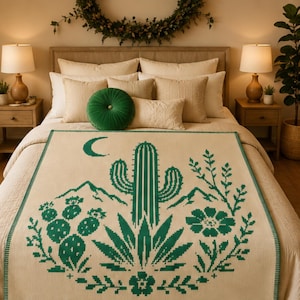May include: A cream-colored woven throw blanket featuring a dark green pixelated desert landscape design. The pattern includes a central saguaro cactus, a crescent moon, mountains, prickly pear cacti, and various desert flora across the surface.