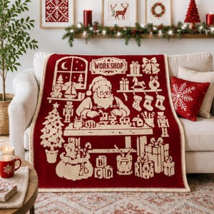 May include: A deep red and cream-colored knit throw blanket featuring a detailed graphic of Santa Claus in a workshop. The design includes the text WORKSHOP, a Christmas tree, toys, stockings, gift boxes, and alphabet blocks in a repeating pattern.