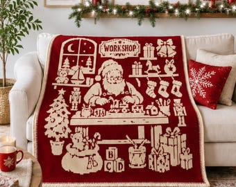 Puede incluir: Una manta de punto en color rojo oscuro y crema con un gráfico detallado de Santa Claus en su taller. El diseño incluye el texto WORKSHOP, un árbol de Navidad, juguetes, calcetines navideños, cajas de regalo y bloques con letras.