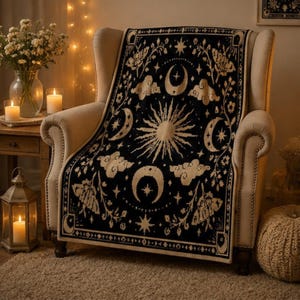 Puede incluir: Una manta tejida en color negro y crema con un motivo central de sol rodeado de lunas crecientes, estrellas, nubes e ilustraciones de polillas. El tejido rectangular tiene un borde decorativo y cubre un sill&oacute;n tapizado en beige.