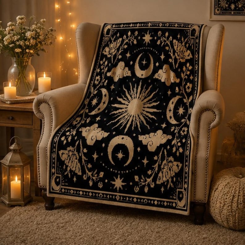 Puede incluir: Una manta tejida en color negro y crema con un motivo central de sol rodeado de lunas crecientes, estrellas, nubes e ilustraciones de polillas. El tejido rectangular tiene un borde decorativo y cubre un sill&oacute;n tapizado en beige.