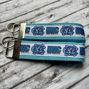 May include: Two light blue keychains with silver key rings. The keychains have a white ribbon with the text "UNC CAROLINA TAR HEELS" and a logo in light blue. The keychains are on a light wood surface.