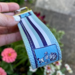 May include: A light blue and white striped keychain wristlet with a gold-colored metal clasp. The wristlet features a small rectangular patch with a cartoon mascot. The background includes pink and white flowers.