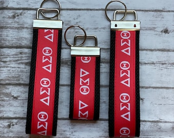 May include: Three fabric key fobs with silver-tone metal hardware and split rings. Each features a central red ribbon with the white Greek letters Delta Sigma Theta, bordered by black textured fabric on both sides of the ribbon.