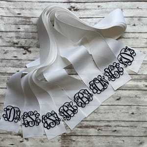 K&ouml;nnte beinhalten: Eine Sammlung wei&szlig;er Satinb&auml;nder mit schwarzen, gestickten Monogrammen in einer dekorativen Schreibschrift. Jedes Band zeigt ein einzigartiges Design aus drei Initialen, wobei die B&auml;nder &uuml;berlappend angeordnet sind, um die verschiedenen Stile zu zeigen.