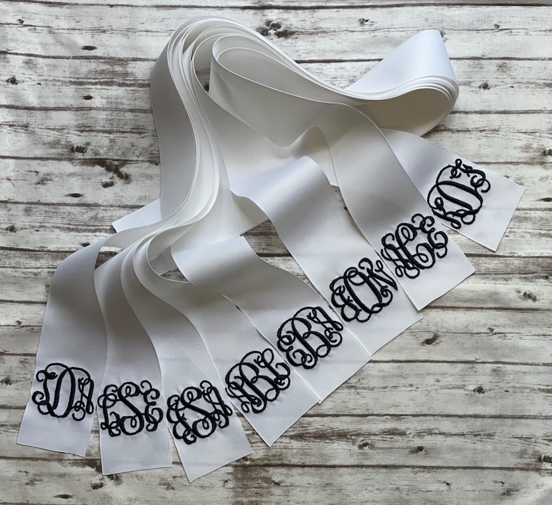 K&ouml;nnte beinhalten: Eine Sammlung wei&szlig;er Satinb&auml;nder mit schwarzen, gestickten Monogrammen in einer dekorativen Schreibschrift. Jedes Band zeigt ein einzigartiges Design aus drei Initialen, wobei die B&auml;nder &uuml;berlappend angeordnet sind, um die verschiedenen Stile zu zeigen.