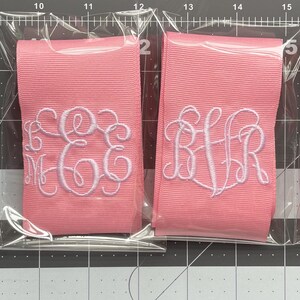 K&ouml;nnte beinhalten: Vier gefaltete Stoffb&auml;nder in durchsichtigen Kunststoffh&uuml;llen. Ein wei&szlig;es Band hat ein rosa gesticktes Monogramm, w&auml;hrend drei rosa B&auml;nder wei&szlig;e gestickte Monogramme aufweisen. Die B&auml;nder liegen auf einer schwarzen Schneidematte.