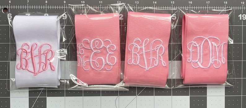 K&ouml;nnte beinhalten: Vier gefaltete Stoffb&auml;nder in durchsichtigen Kunststoffh&uuml;llen. Ein wei&szlig;es Band hat ein rosa gesticktes Monogramm, w&auml;hrend drei rosa B&auml;nder wei&szlig;e gestickte Monogramme aufweisen. Die B&auml;nder liegen auf einer schwarzen Schneidematte.