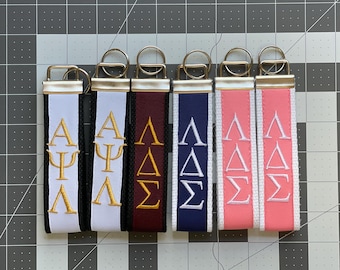 May include: Six fabric key fobs with gold-tone metal hardware and split rings. The fobs feature embroidered Greek letters in various color combinations: white with gold, black with gold, maroon with gold, navy with white, and pink with white.