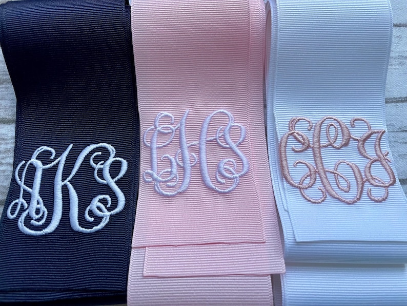 K&ouml;nnte beinhalten: Drei Rollen Ripsband in Marineblau, Hellrosa und Wei&szlig;. Jedes Band ist mit einem dekorativen, gestickten Monogramm in Schreibschrift versehen. Das marineblaue Band hat wei&szlig;e Stickereien, das rosa Band hat wei&szlig;e Stickereien und das wei&szlig;e Band hat ros&eacute;goldene Stickereien.