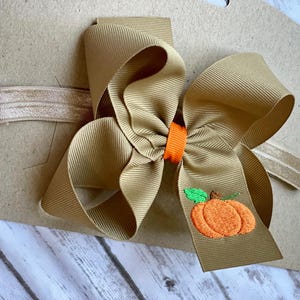 K&ouml;nnte beinhalten: Ein braunes Stoffhaarband mit einer gro&szlig;en braunen Schleife und einem gestickten orangefarbenen K&uuml;rbis mit gr&uuml;nem Stiel.