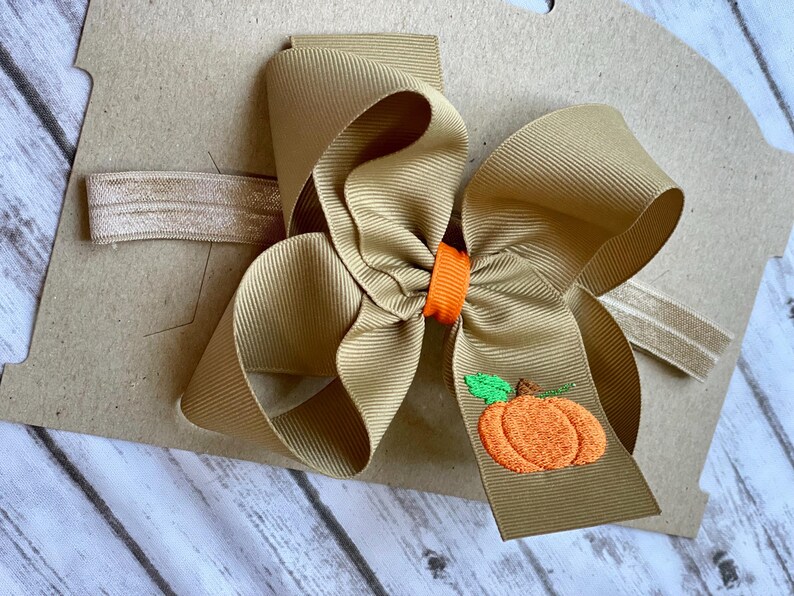 K&ouml;nnte beinhalten: Ein braunes Stoffhaarband mit einer gro&szlig;en braunen Schleife und einem gestickten orangefarbenen K&uuml;rbis mit gr&uuml;nem Stiel.