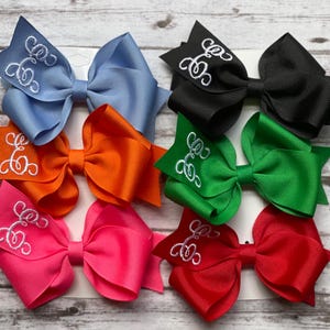 May include: A collection of eight colorful hair bows in various shades, including blue, orange, pink, black, green, and red. Each bow features a white embroidered monogram. The bows are arranged on a light wood-grain surface.