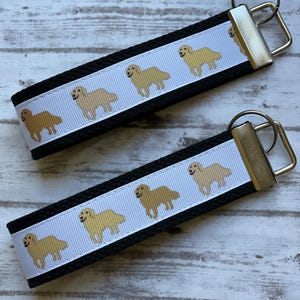 May include: Two black keychains with a white ribbon featuring a repeating pattern of golden retriever dogs. The keychains have silver-coloured metal hardware and are displayed on a light wood-grain surface.